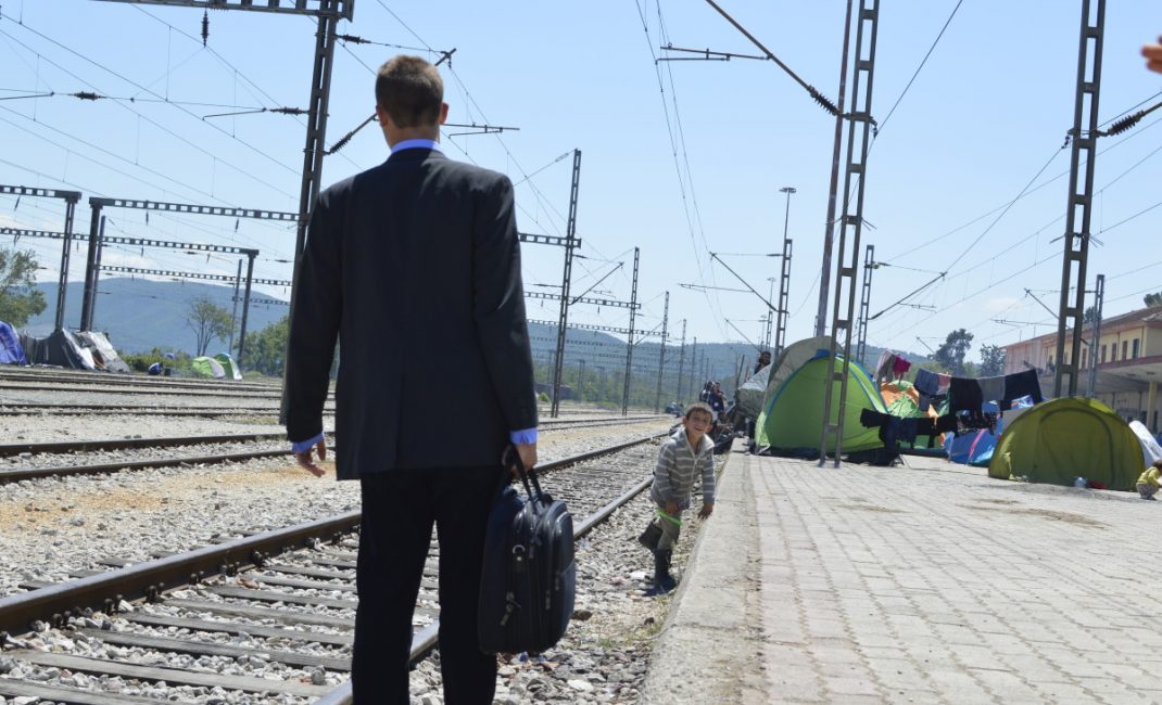 A kid looks curiously at a dressed-up man with an overnight bag on the rail of the Idomeni station
