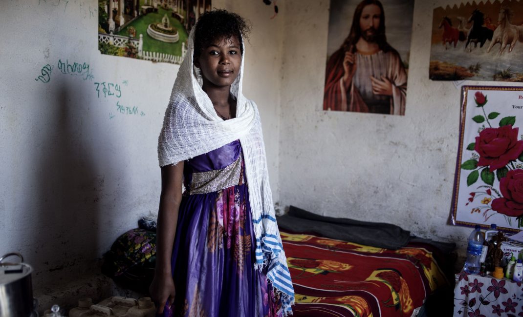 Teenage girl wearing purple dress and white head scarf, smiling in front of a poster of Jesus.