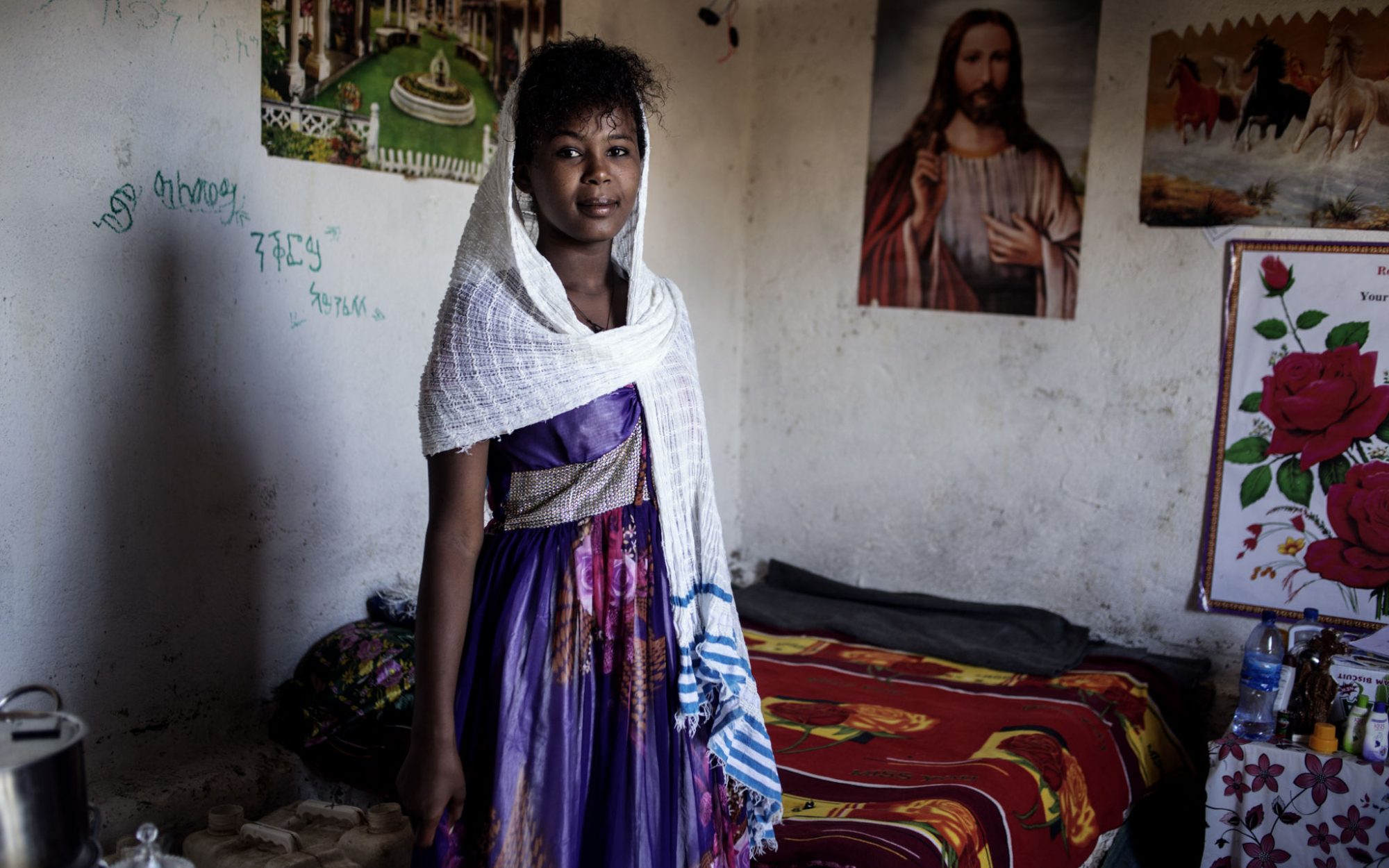 Teenage girl wearing purple dress and white head scarf, smiling in front of a poster of Jesus.