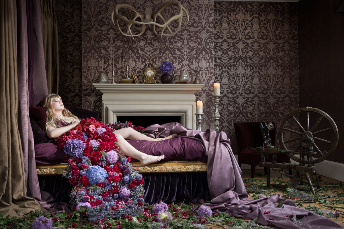 A woman lies peacefully sleeping on a purple bed, beneath a blanket of flowers.