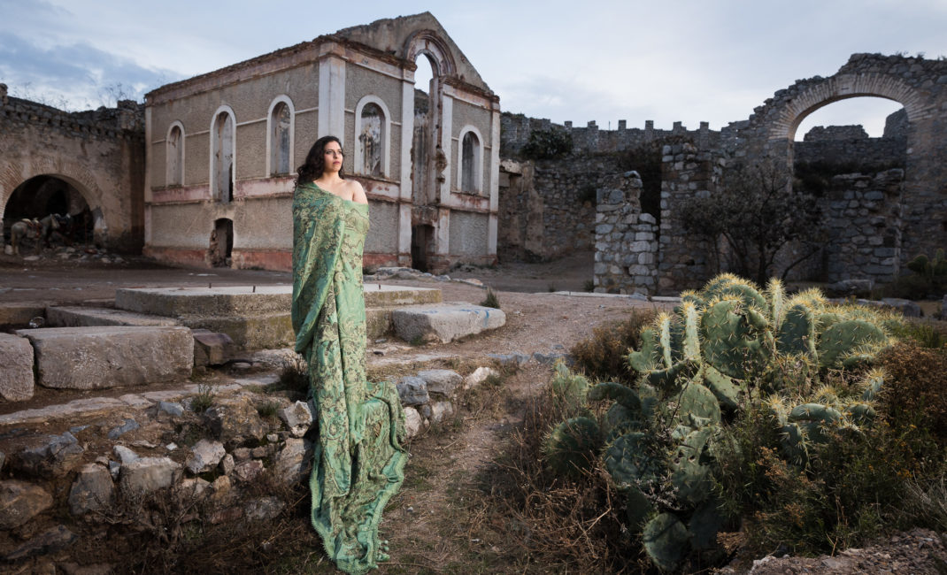 A woman stands, wrapped in green fabric, next to a crop of cacti and in front of ruins.