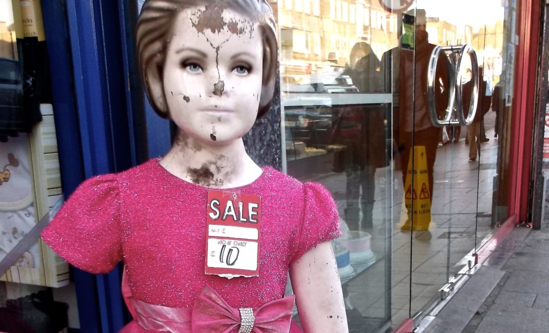A defaced mannequin girl stands outside a shop, wearing a pink dress and a pricetag.