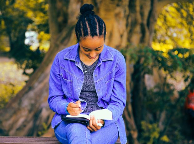 Woman wearing denim sit on a bench in front of a tree engrossed in a book.
