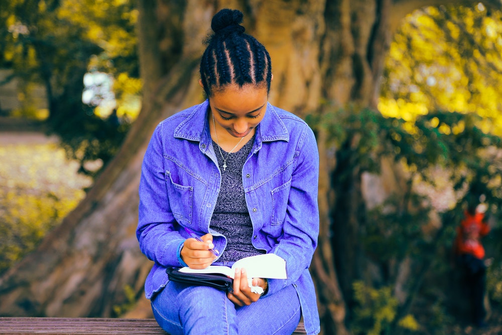 Woman wearing denim sit on a bench in front of a tree engrossed in a book.