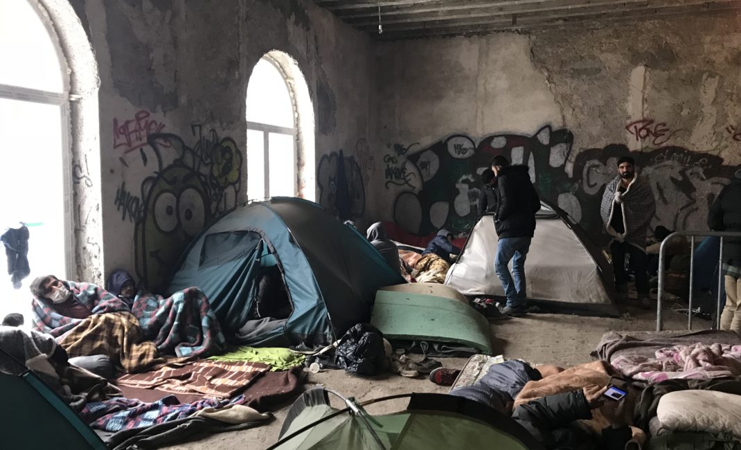 People huddle in blankets as tents, mattresses and rugs fill a bare, crumbling, graffiti-covered room.