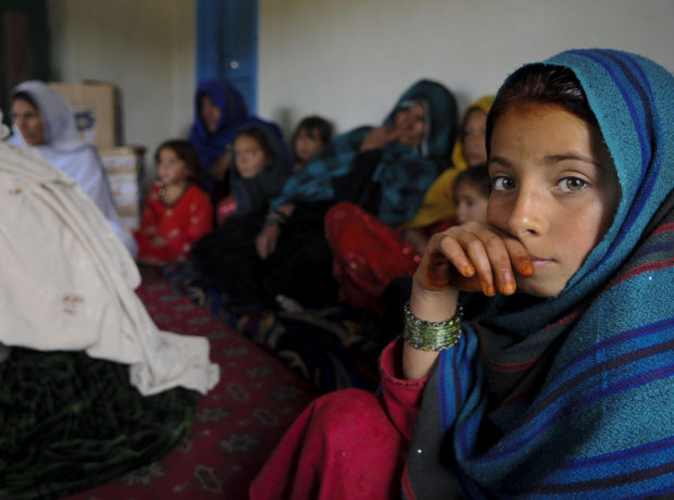 A young Afghan girl in a blue veil looks directly at the camera while a group of Afghan women and girls sit behind her out of focus