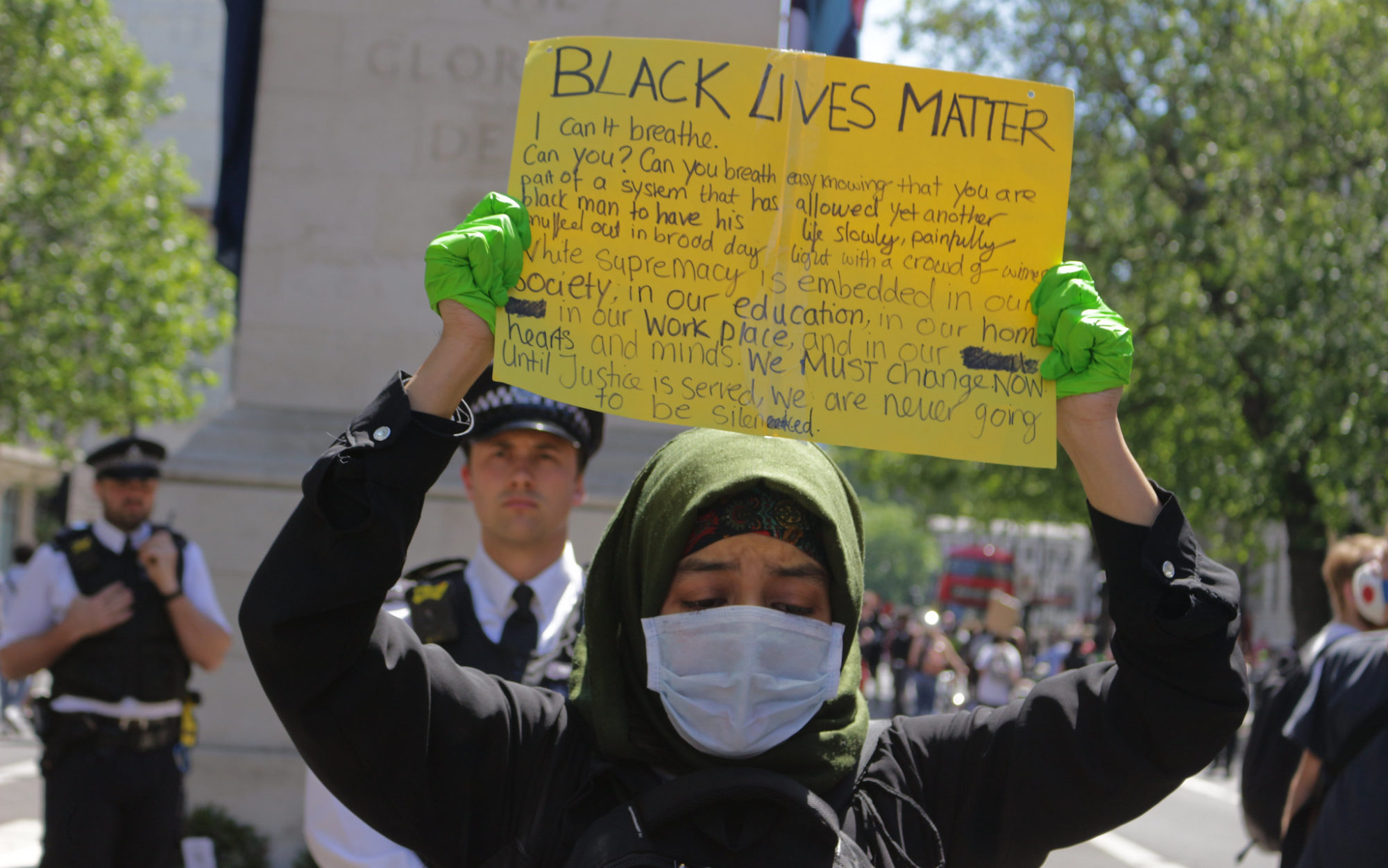 Black Lives Matter protest in London May 2020, a young Muslim woman, wearing a hijab, a face mask and green surgical gloves, looks down as she lifts a homemade BLM placard