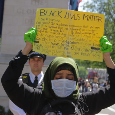 Black Lives Matter protest in London May 2020, a young Muslim woman, wearing a hijab, a face mask and green surgical gloves, looks down as she lifts a homemade BLM placard