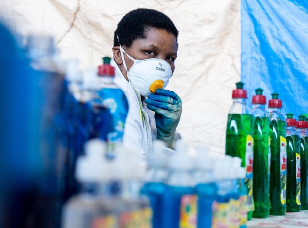 A woman wearing a face mask, gloves and white coat, stands behind detergents and hand sanitisers