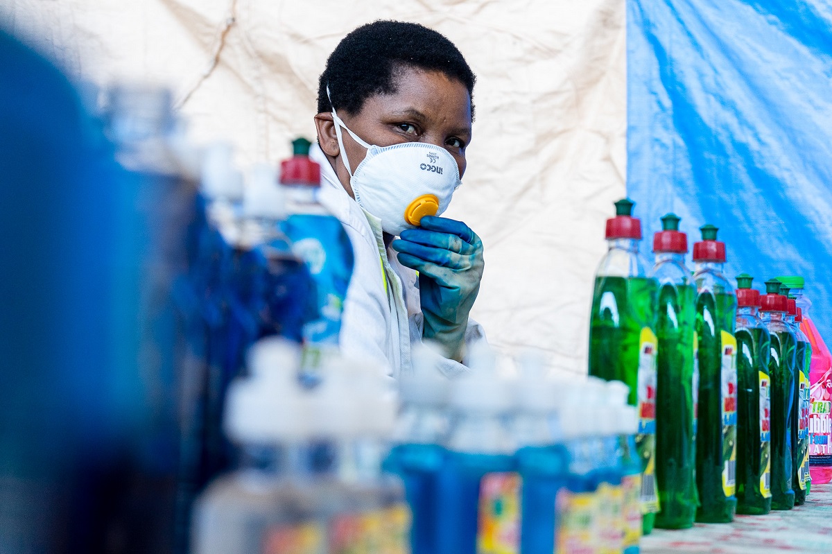 A woman wearing a face mask, gloves and white coat, stands behind detergents and hand sanitisers