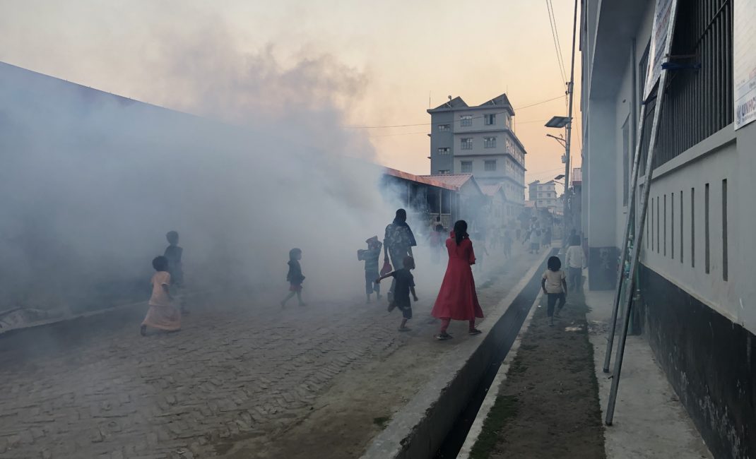 Rohingya refugees, adults and children, walk along a brick road through thick clouds of mosquito repellent in Bhasan Char.