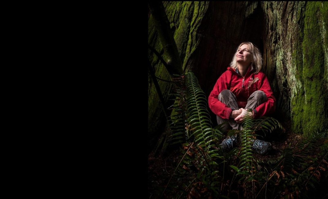 Ecologist Suzanne Simard photographed in Stanley Park, sitting, legs crossed, at the base of a large tree and looking up, smiling.