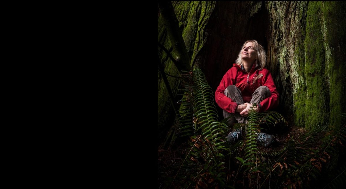 Ecologist Suzanne Simard photographed in Stanley Park, sitting, legs crossed, at the base of a large tree and looking up, smiling.