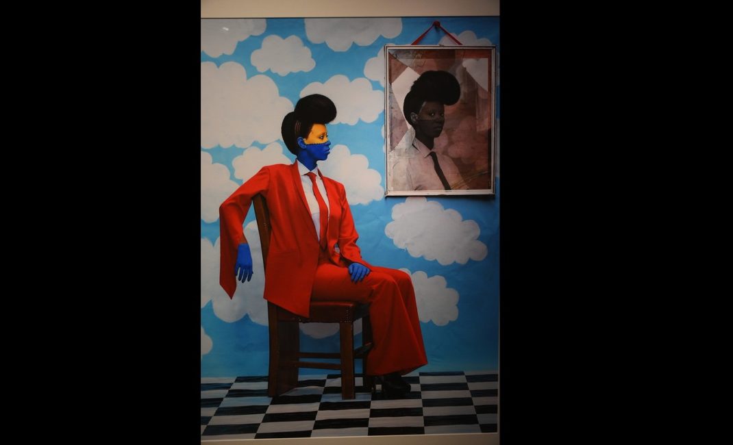 A woman painted blue and yellow, wearing a bright red trouser suit, sits on a black and white tiled floor in front of wallpaper of blue sky and white clouds