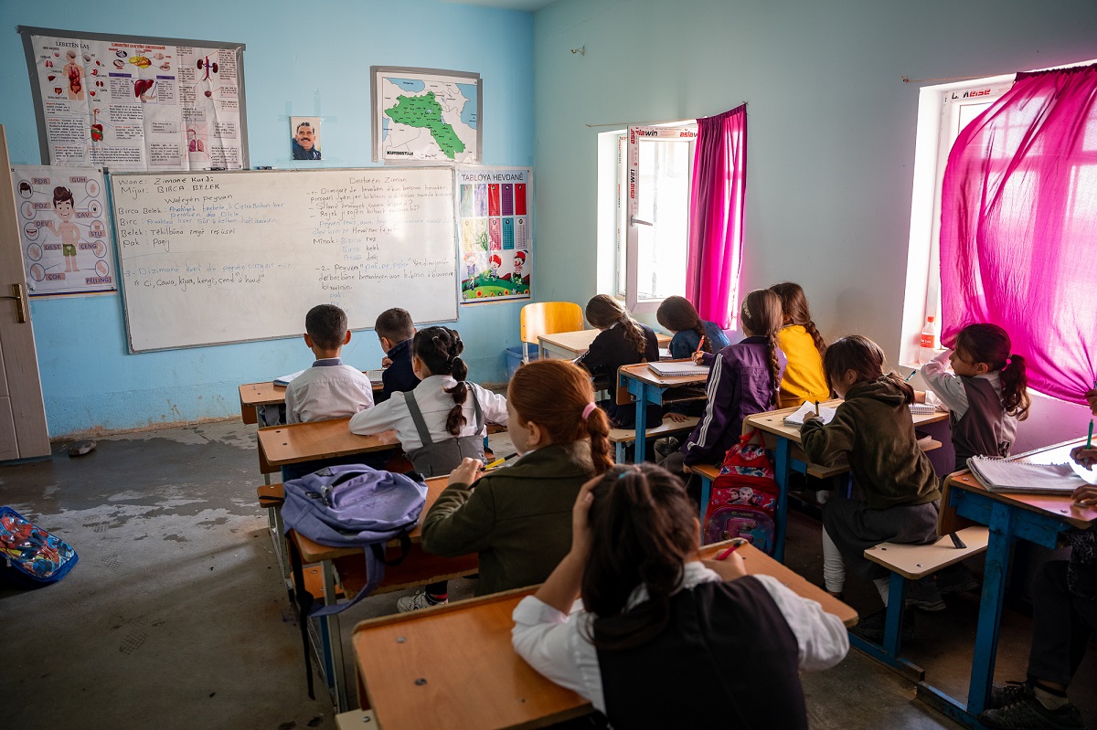 A school in the Makhmour refugee camp. Around 10 children are sitting on desks, looking at the board.