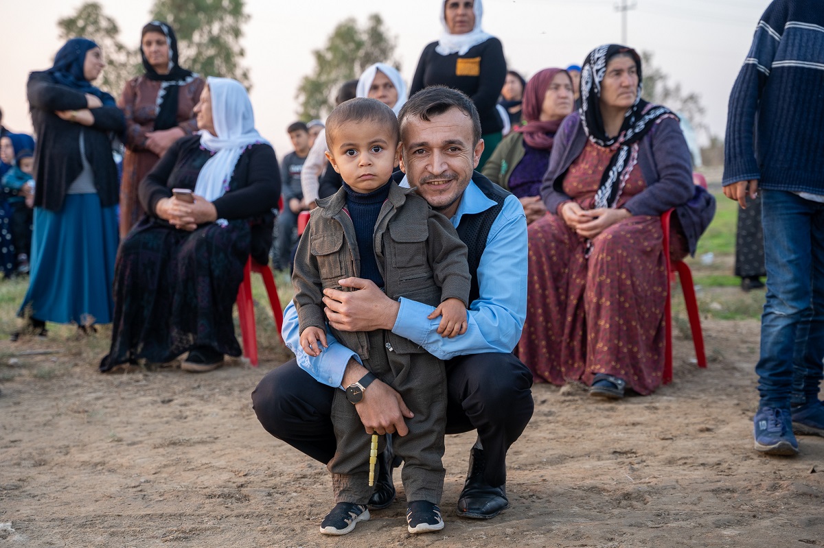 A middle aged man and a boy standing, the man is hugging the child from behind, the boy is wearing traditional Kurdish clothing