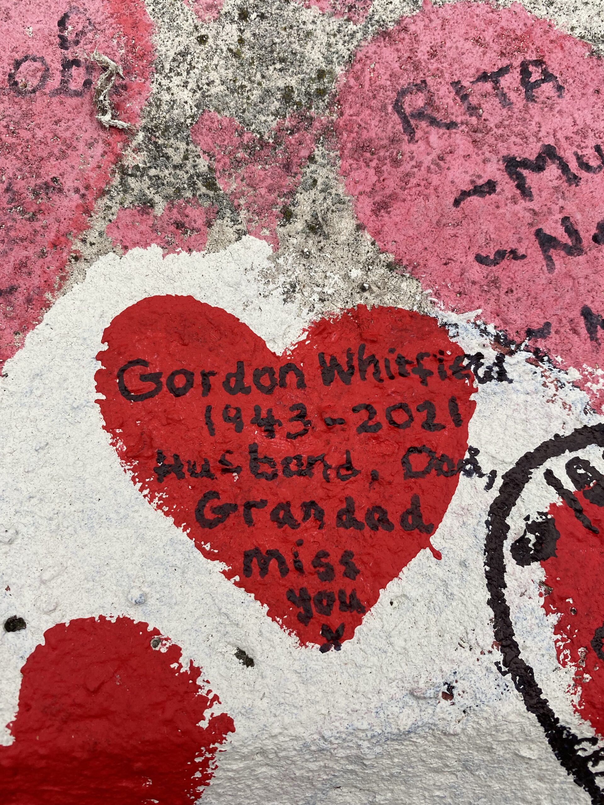 Close-up photo of the COVID-19 Memorial Wall with a red heart in Gordon Whitfield's memory. The rest of the wall is covered by red and pink hearts along with the names of deceased loved ones