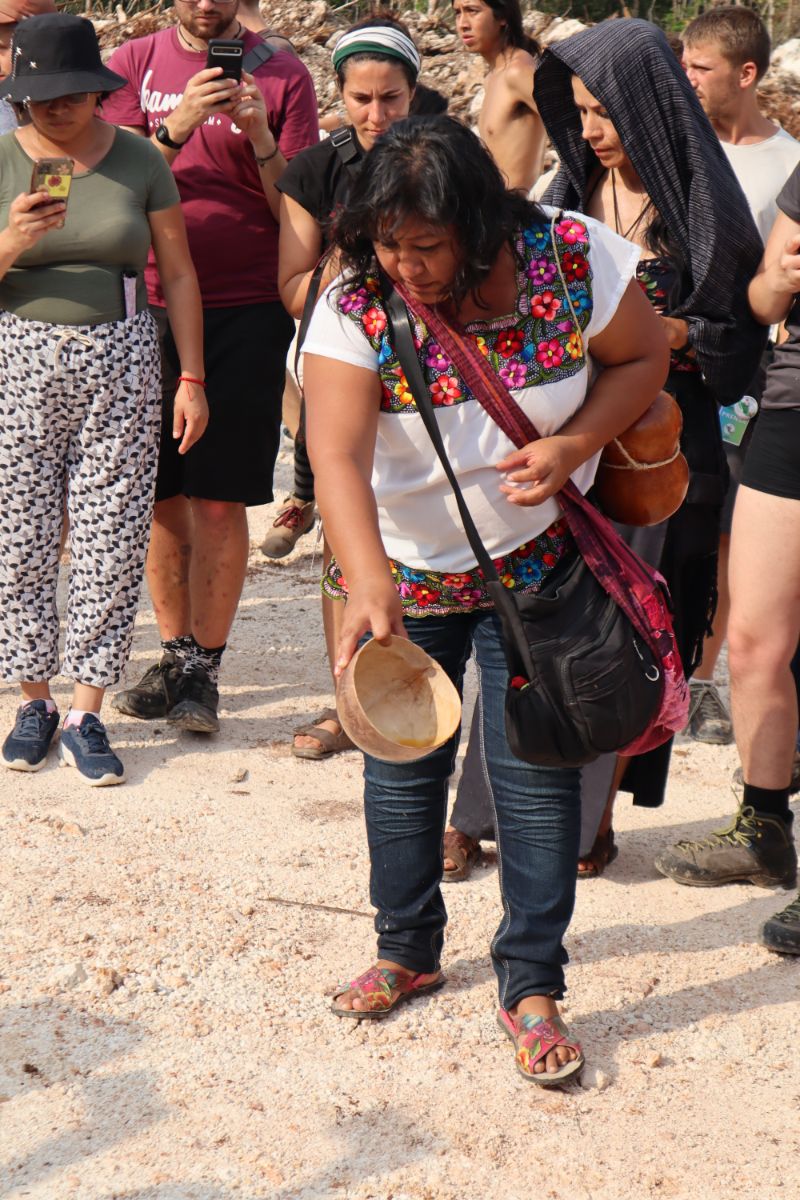 A woman surrounded by spectators stands, emptying a container over the land beneath her feet
