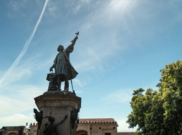 Statue of colonial figure Christopher Columbus towering over a building and trees while pointing upwards to the sky in Santa Domingo