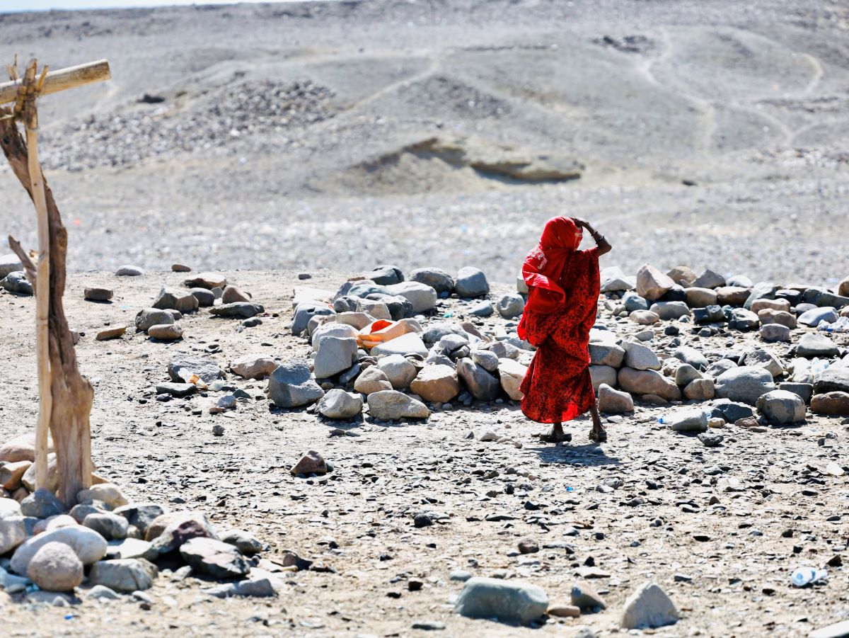 Biruk, a woman dressed head-to-toe in bright red garments has her back turned to the camera. She is looking off into the distance. She is in the desert near a circle of rocks