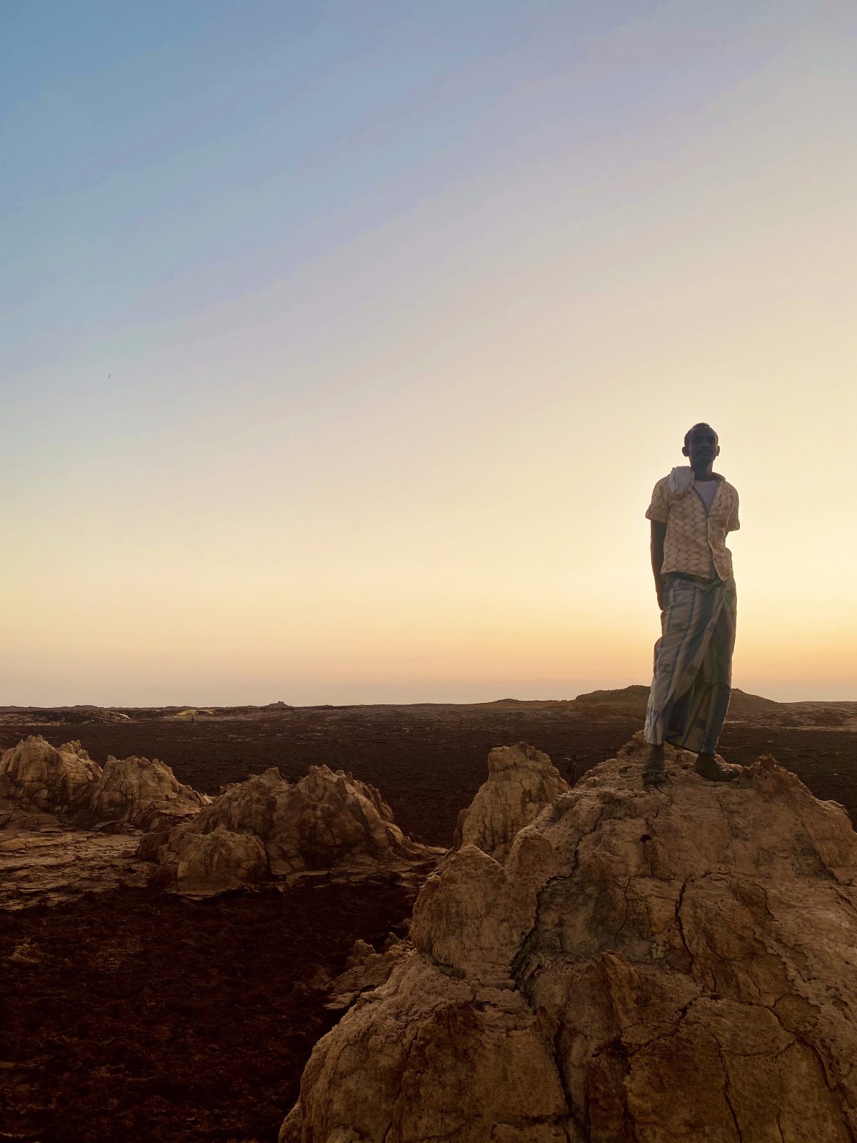 The tribe leader, Aboubakr, stands atop a rock structure in loose clothing. He stands confidently with his hands behind his back as the sun sets behind him