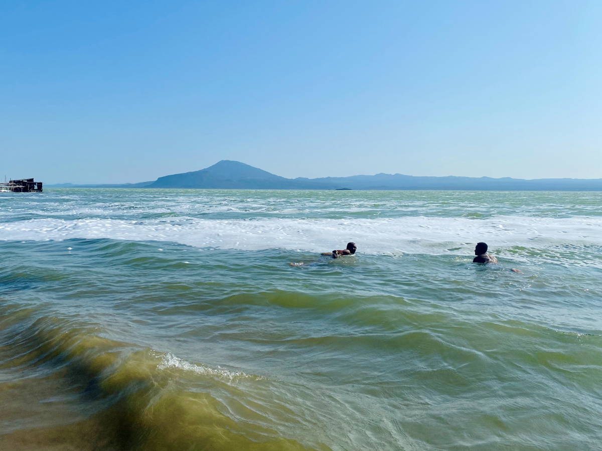 Two men swim in the sea. The water is green and blue with white seafoam. There are mountains in the distance and the sky is blue