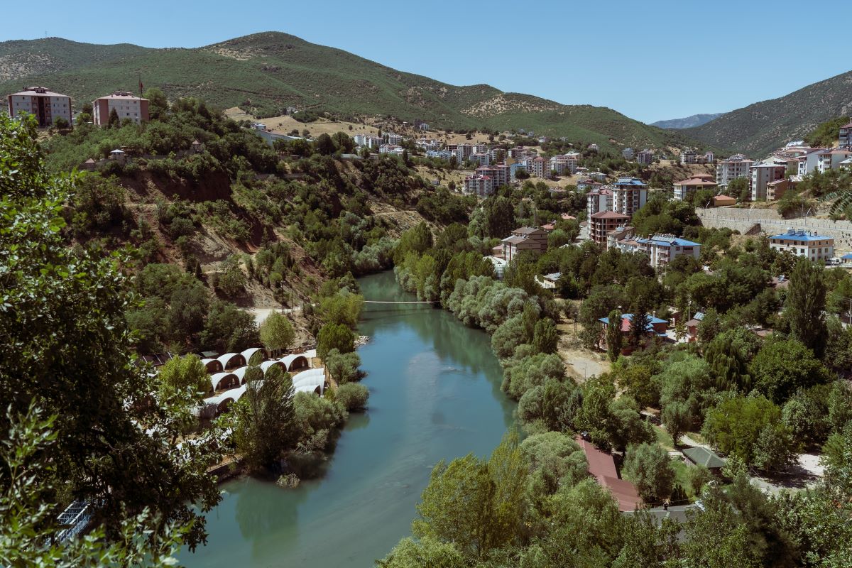 A blue river flows through a town. There are lush green trees which line the water and there are hills in the background