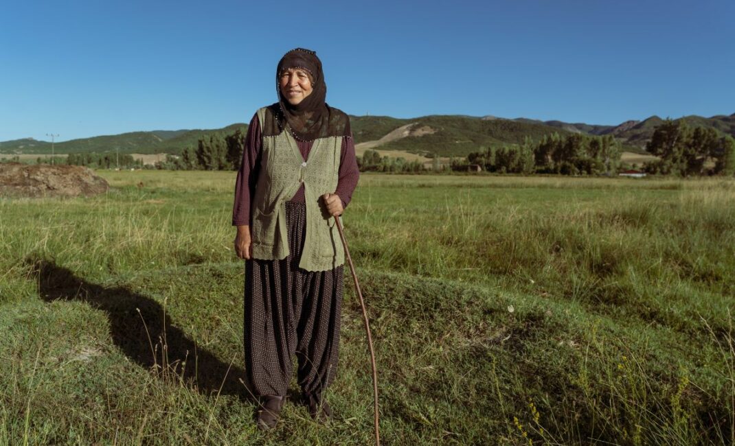 A woman in loose trousers and a dark purple long sleeved top stands in the foreground holding a walking stick. Her hair is covered, she is elderly and smiling. In the background there are hills