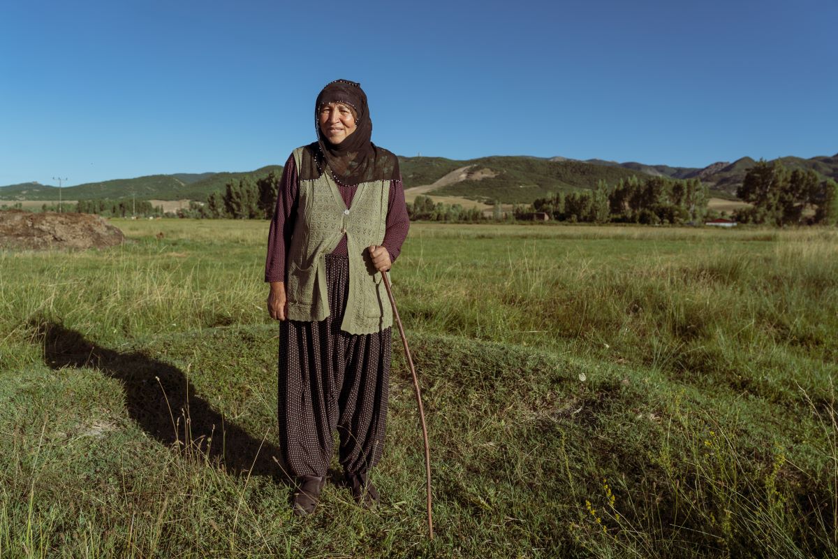 A woman in loose trousers and a dark purple long sleeved top stands in the foreground holding a walking stick. Her hair is covered, she is elderly and smiling. In the background there are hills 