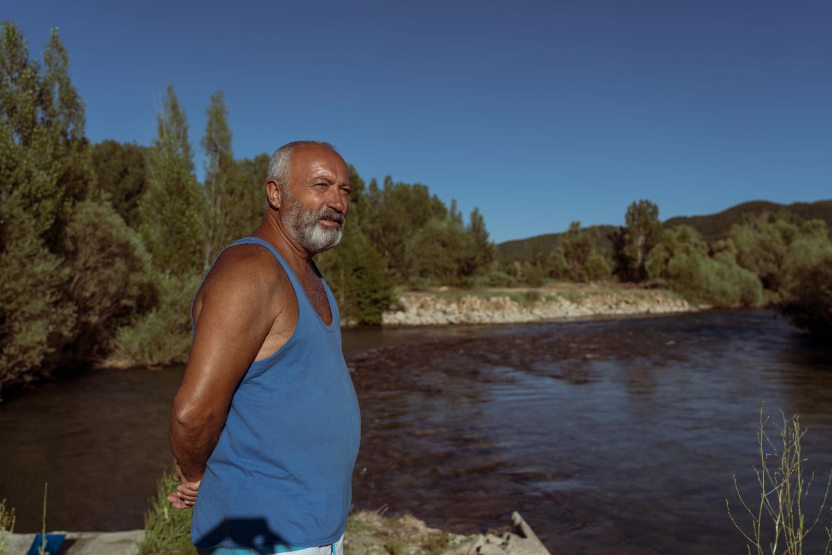 A tanned man in a blue tank top stands with his hands behind his back. He is staring off into the distance. The river flows behind him