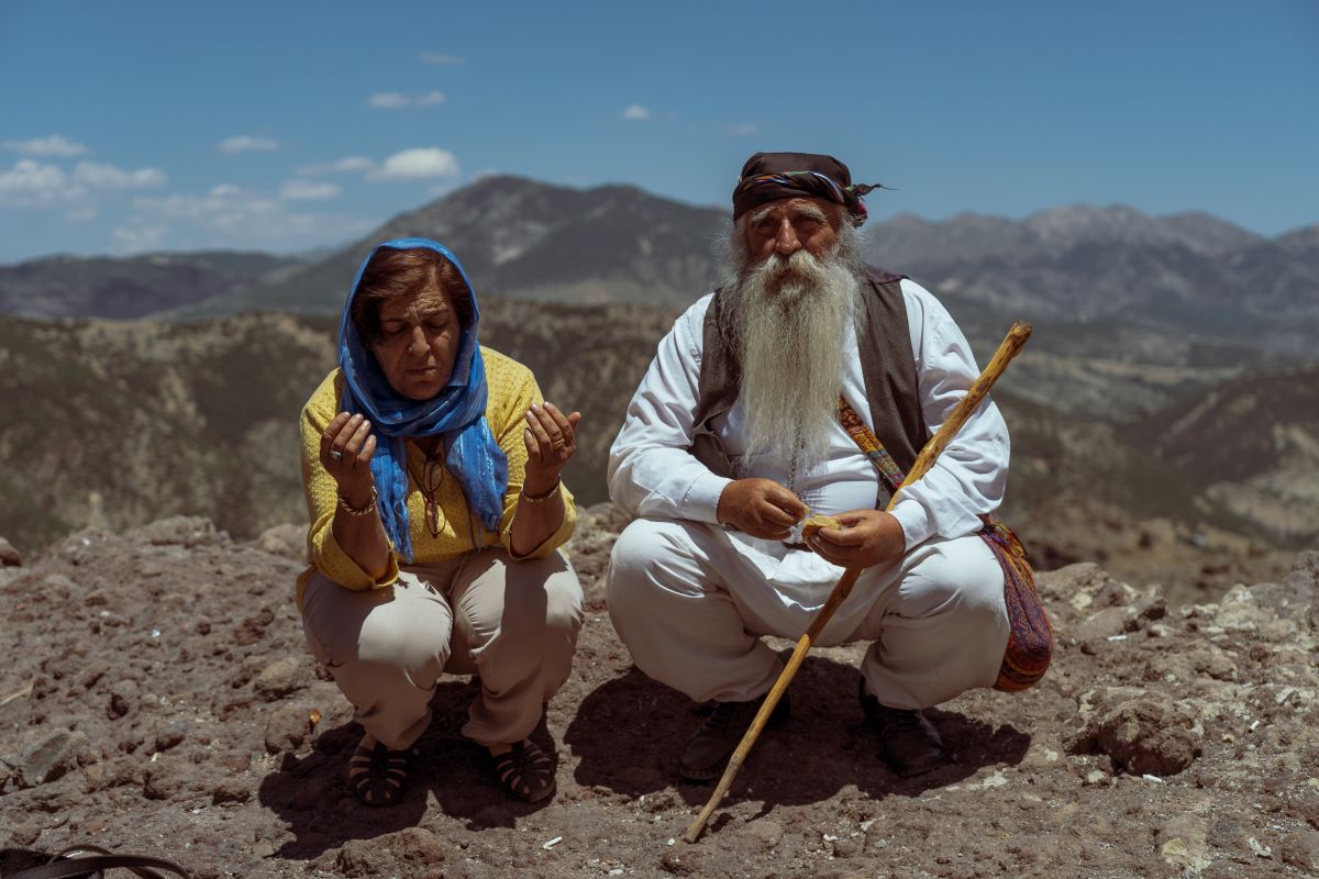 A man dressed in white with a grey vest and head scarf crouches with a woman in a yellow top, blue head scarf, and white trousers. The man is holding a stick. They appear to be atop a hill with mountains in the background