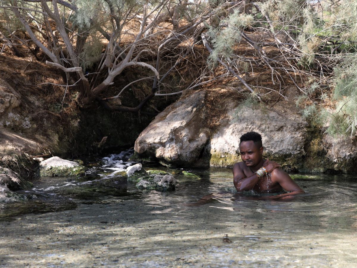 A man bathes in a clear, shallow pond. He is surrounded by dry land and sparse trees. The rocks near the water are mossy 