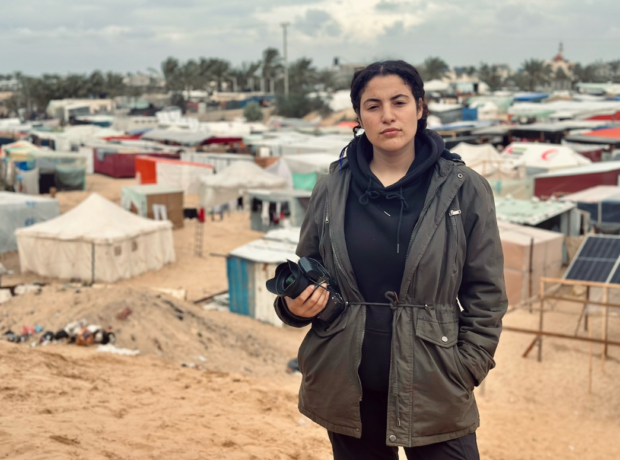Bisan Owda stands in front of a camp holding her camera