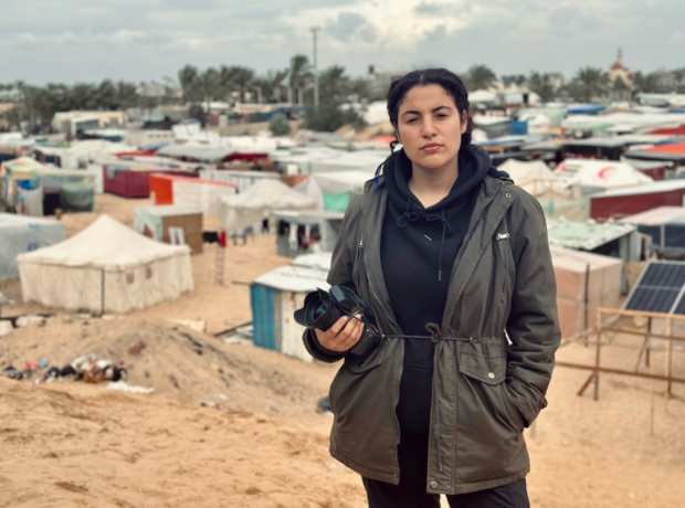 Bisan Owda stands holding a camera with a refugee camp in the background