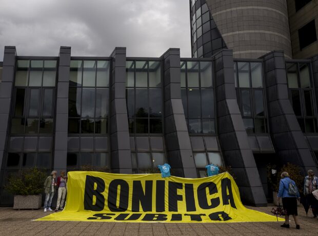 "Bonifica Subito" black text on yellow banner displayed by protesters outside court