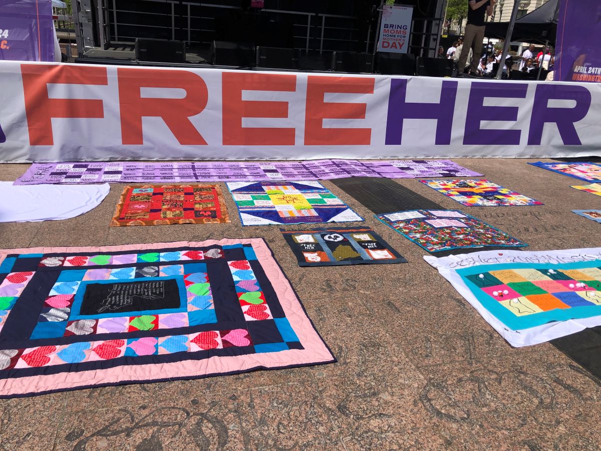 A banner which reads "Free Her" in orange and purple text is hung up before a stage. There are handmade quilts on the ground in front of the banner