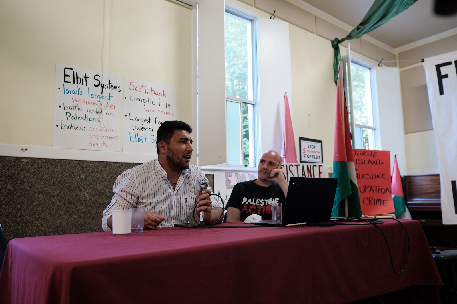 Ronnie Barkan looks intently at Mohammed Al Zaza while Zaza delivers a speech. They are sat at a long table with a red table cloth. There are informative posters about Palestine Action behind them and they are surrounded by a few Palestinian flags.