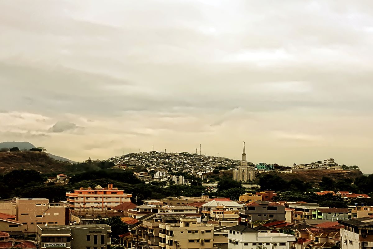 a wide angle view of the city of Guayaquil, with many buildings, a spire, and some hills in the distance