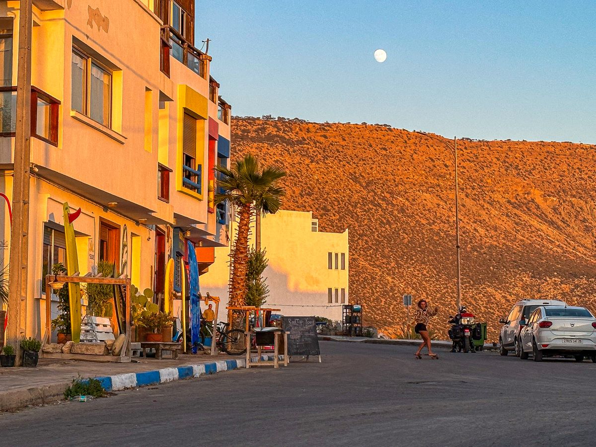 a golden hour photo of a colourful block of flats with the moon in the background. a person is skateboarding along the road parallel