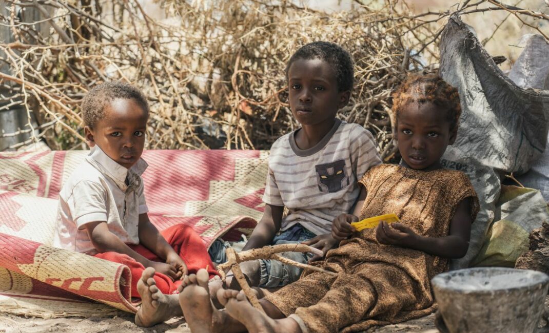 three children sit together in the shade of a shrub