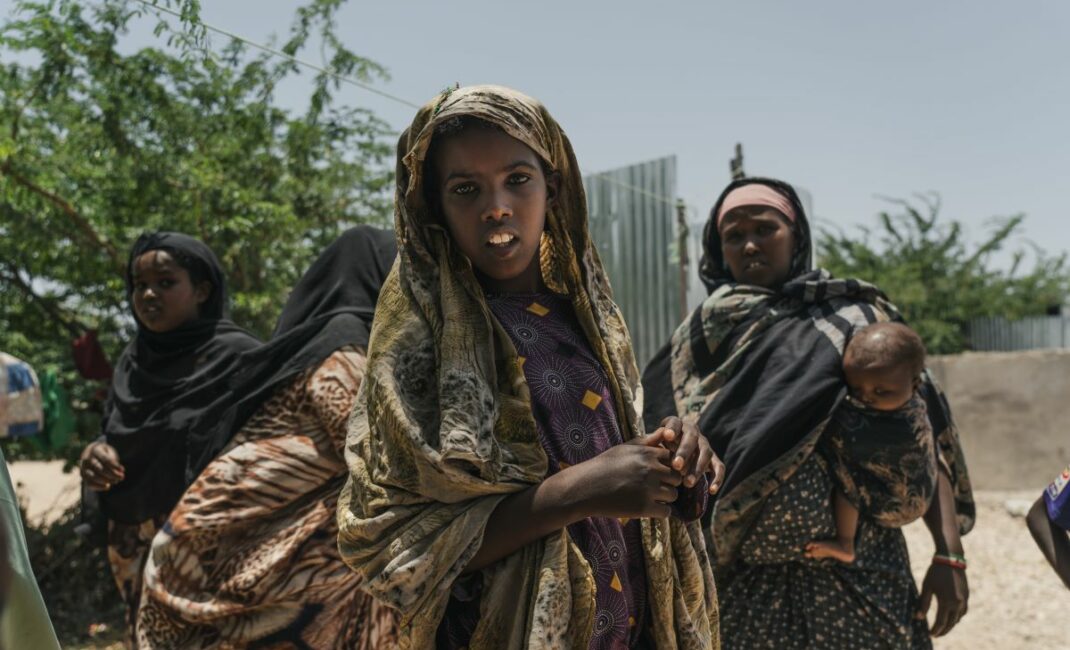 a young veiled girl stares at the camera with some women and girls behind her