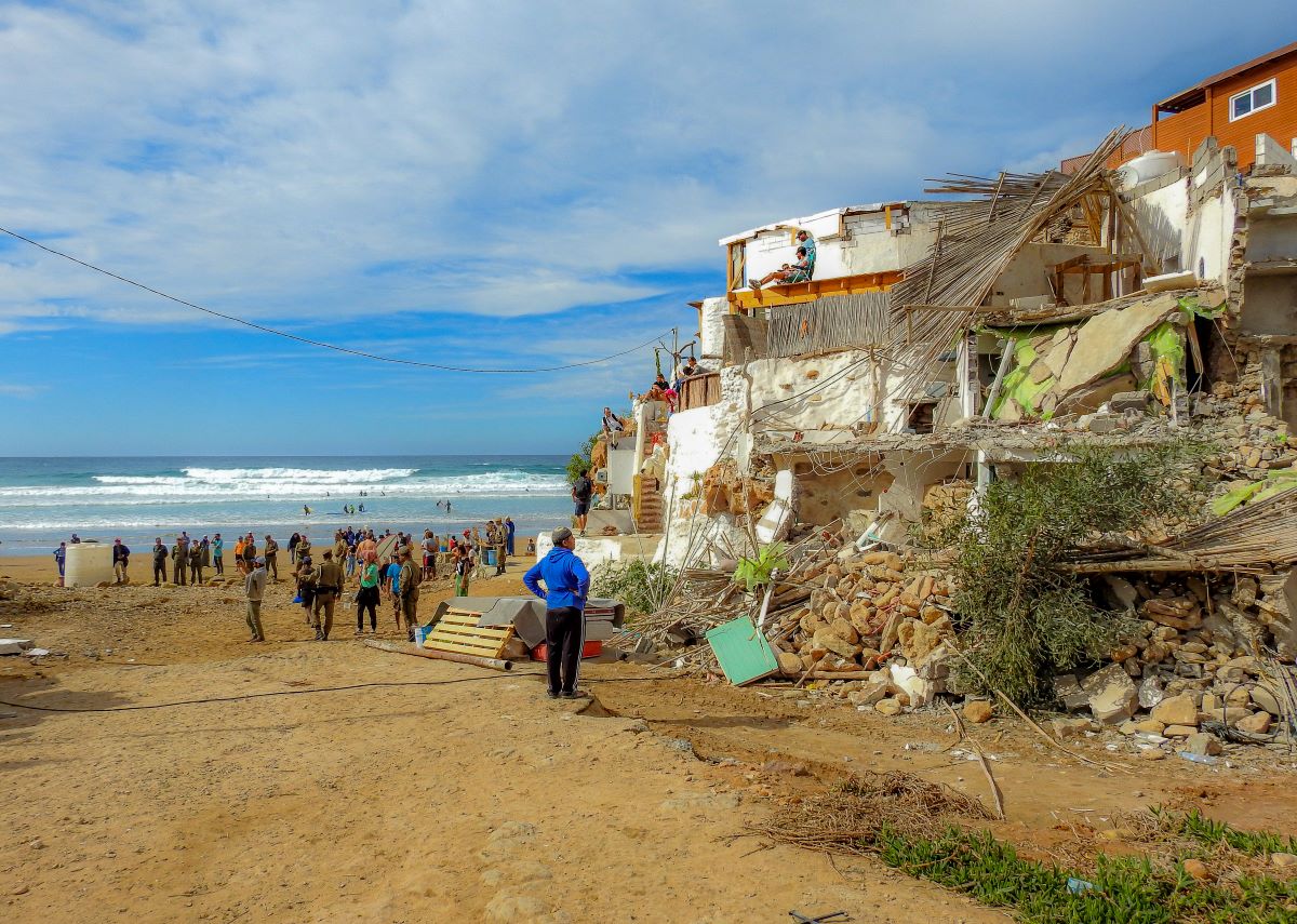 a man looks at the rubble of a house. there is a large group congregating on the beach behind him