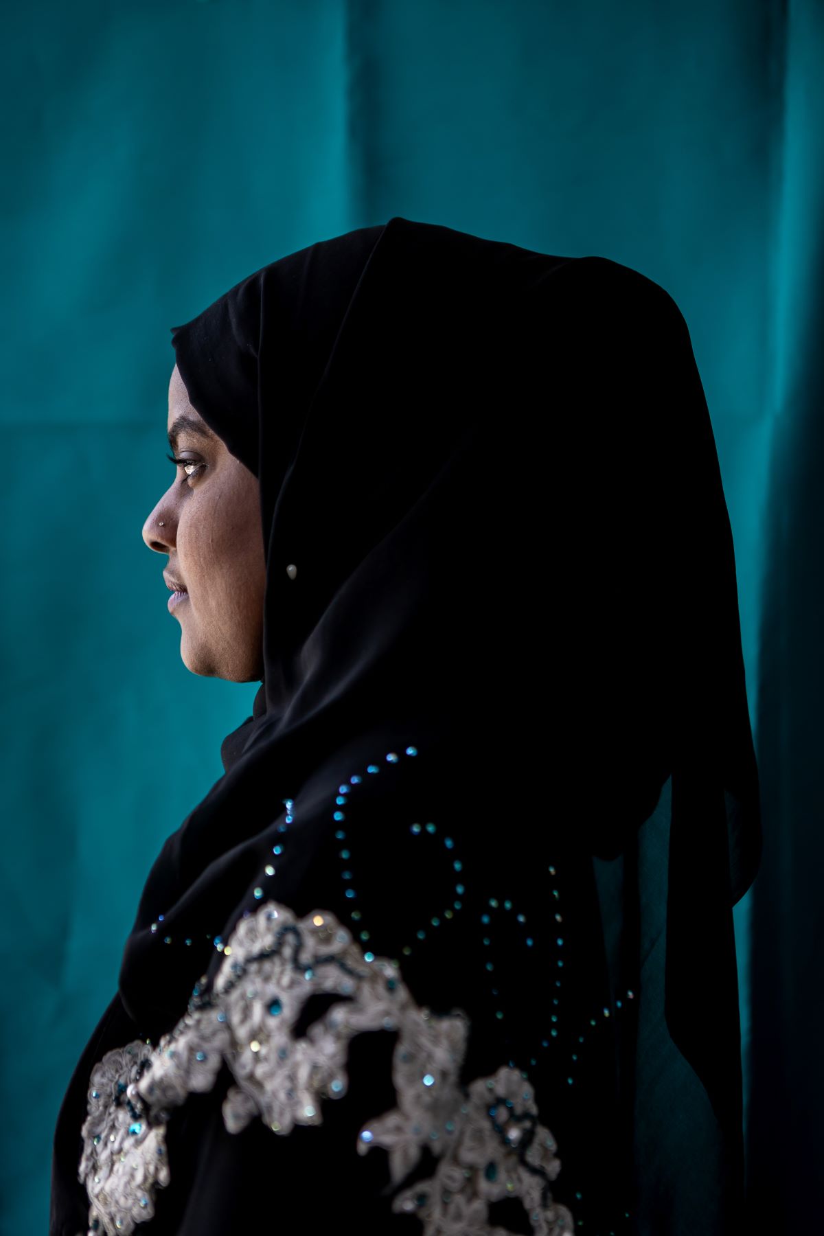 A side profile shot of a woman in a black hijab and an embroidered top. The background behind her is blue