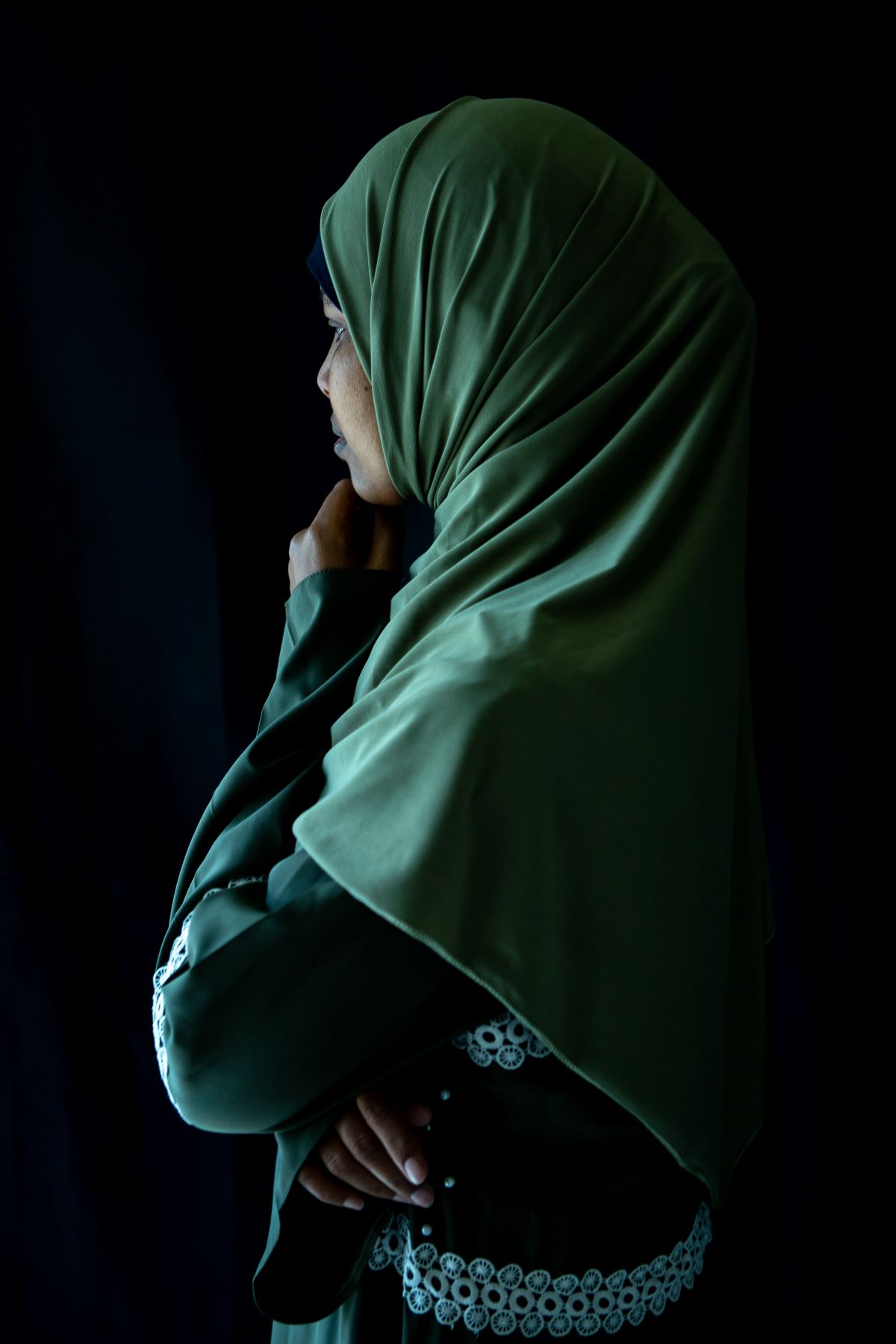 A woman in a green hijab stands looking away from the camera resting her chin on her fist