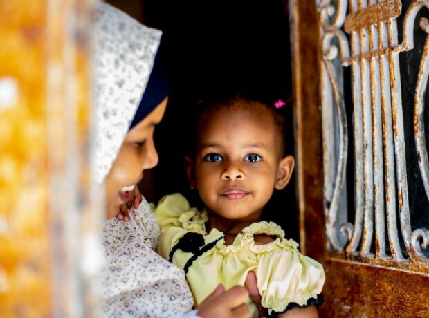 A woman stands in a doorway holding her daughter. She is wearing a white patterned hijab and is smiling at her daughter who looks at the camera. Her daughter has hot pink hair clips in and is wearing a light yellow top