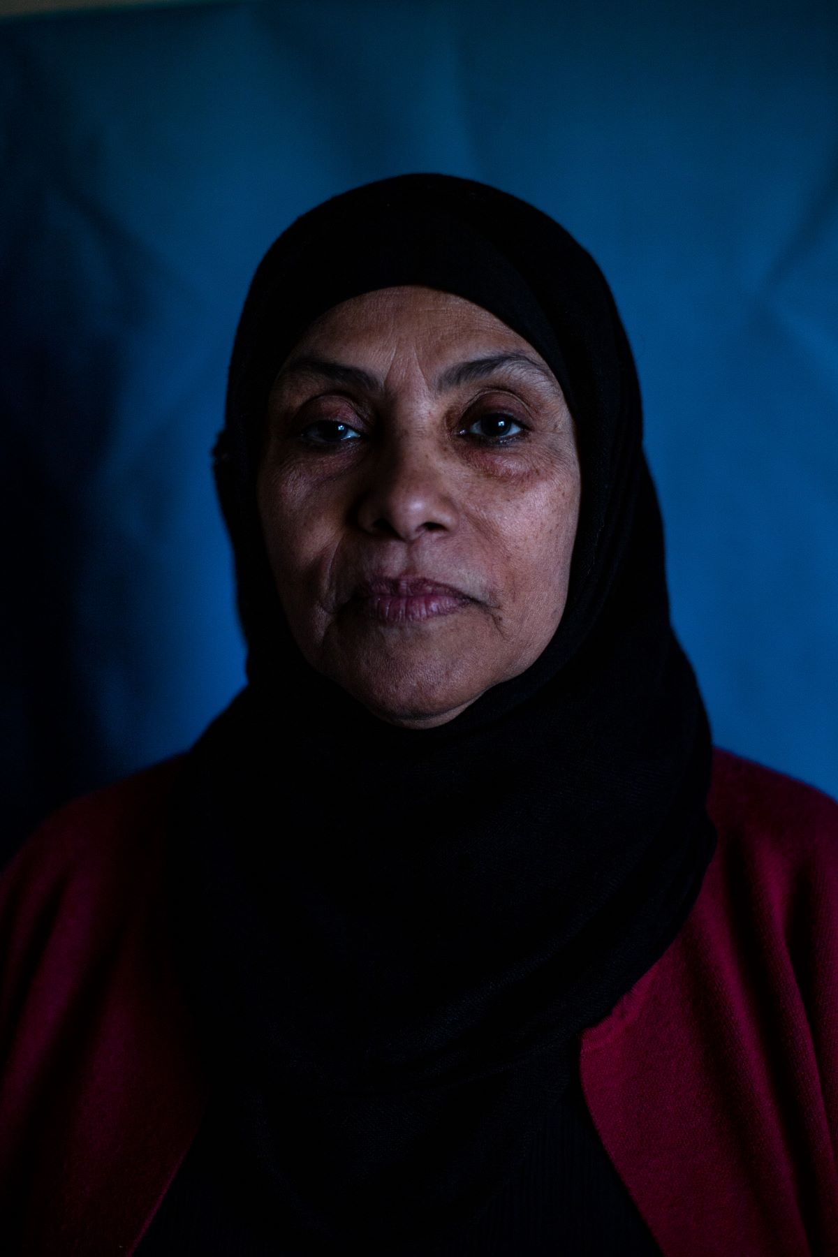 A woman wearing a black hijab and a red cardigan looks at the camera. The lighting is low and the background is blue