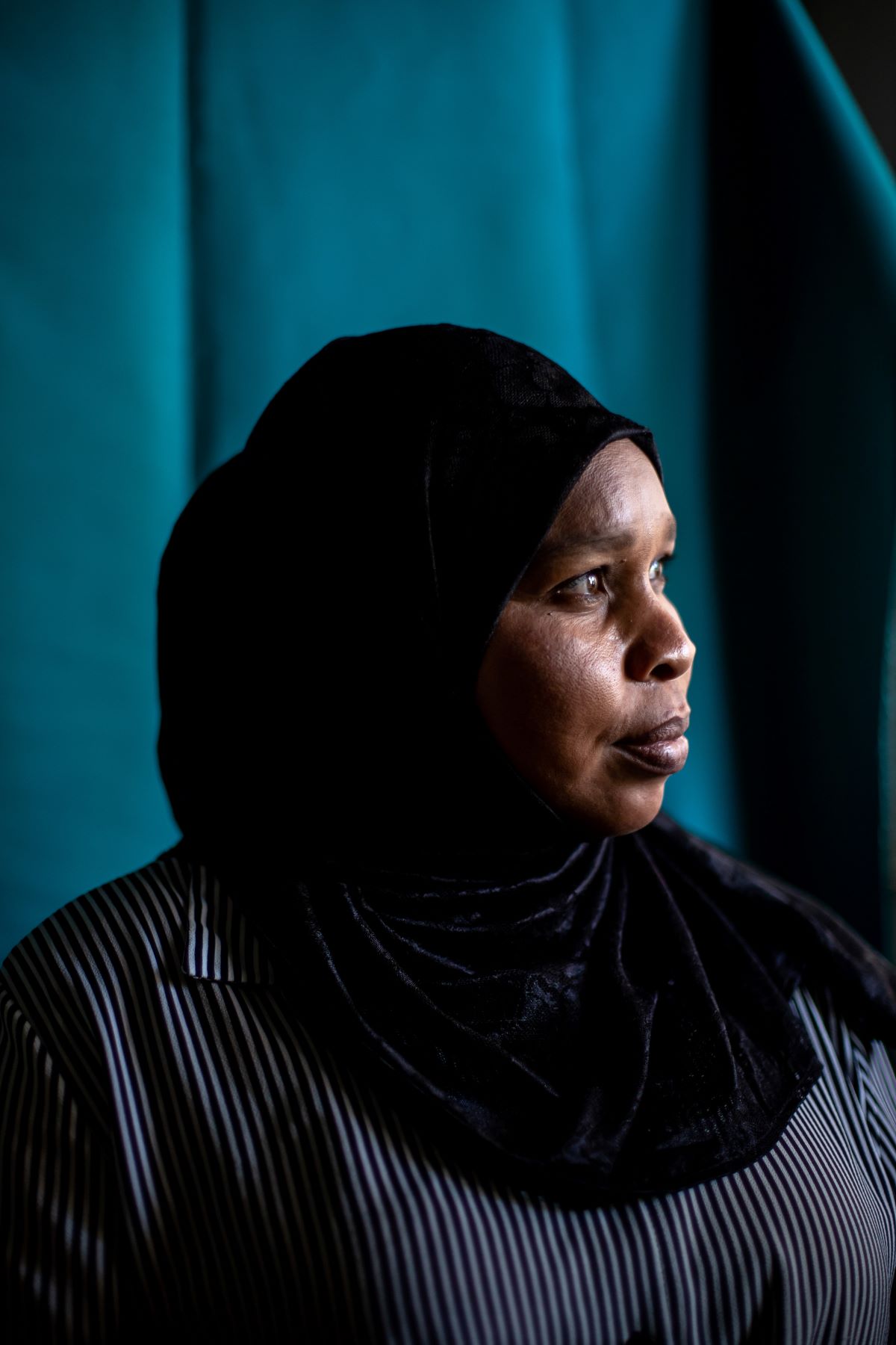 A woma in a black hijab and a pinstriped shirt looks away from the camera to the side. The background is blue