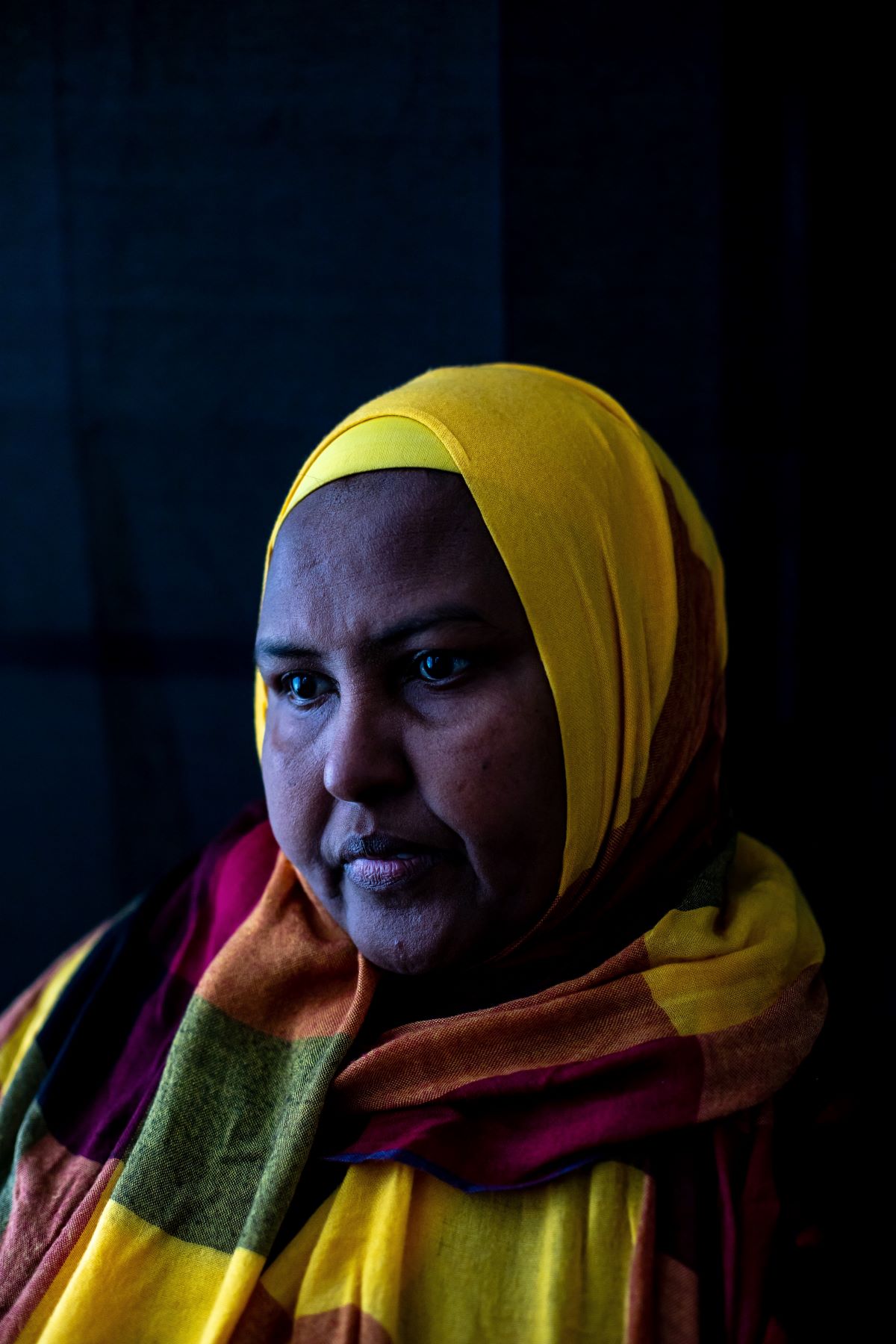 A woman in a multicoloured hijab looks down and away from the camera. Her hijab checked with yellow, green, pink, and orange. The lighting is giving off a blue colour that reflects on her face