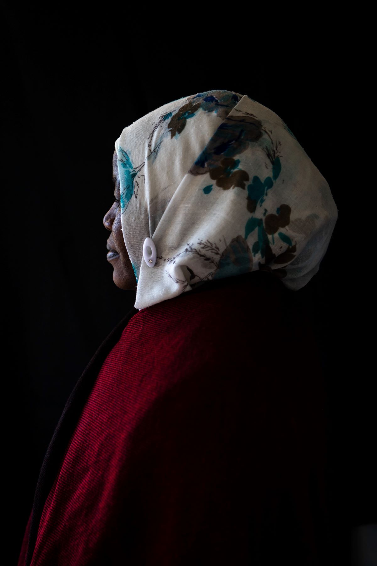 A woman in a white patterned hijab and a red top looks away from the camera. The background is black