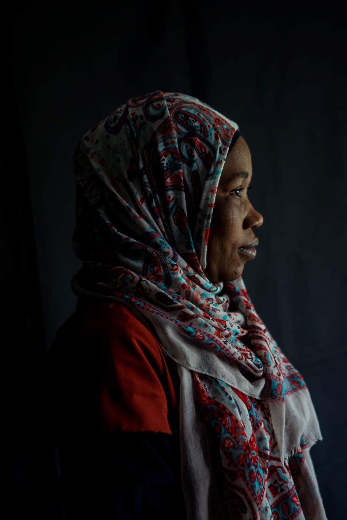 A side profile of a woman wearing a grey, red, and blue hijab. The background is black